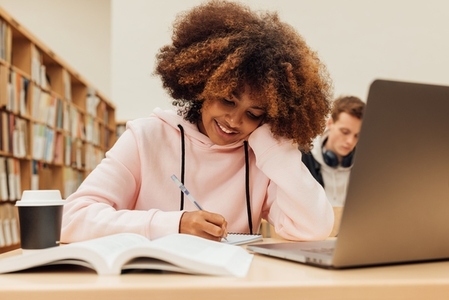 Smiling student with curly hair writing on notebook while sitting at desk in library
