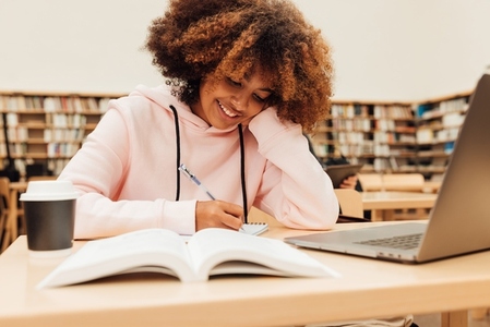 Smiling student writing in library  Girl with curly hair sitting at desk and learning