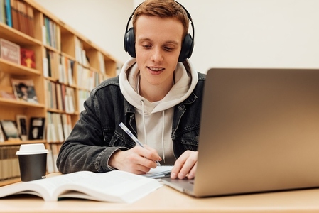 Smiling guy in headphones sitting at desk in library and writing