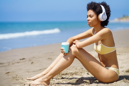 Black woman in headphones drinking takeaway beverage while relaxing on beach