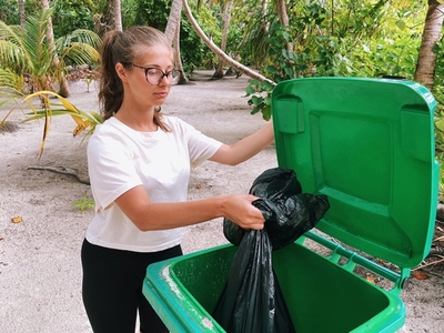 Young woman putting big black bag with garbage in green trash bin on tropical island