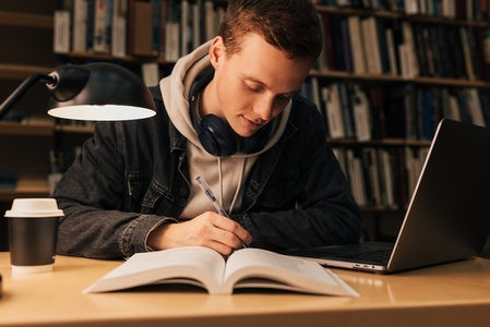 Young man writing while sitting at desk with laptop in library
