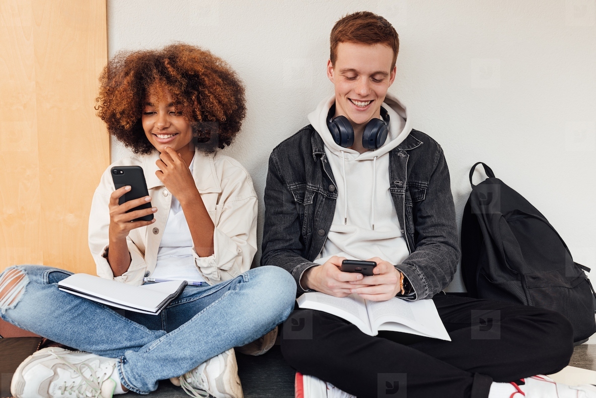 Two smiling classmates sitting at wall and using their smartphones
