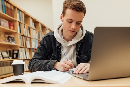Male student writing while sitting at desk in college library