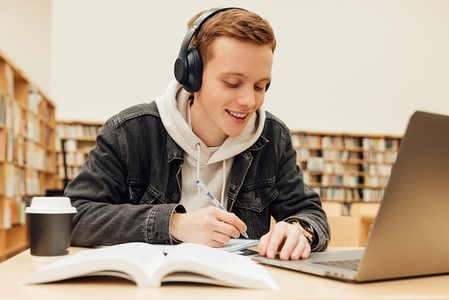Smiling college student wearing wireless headphones while writing on exercise book in library  Guy with ginger hair doing homework