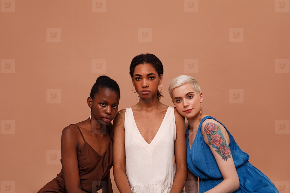 Three beautiful women with different skin color looking at camera. Young females in dresses sitting on brown backdrop