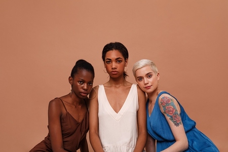 Three beautiful women with different skin color looking at camera  Young females in dresses sitting on brown backdrop