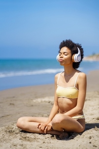 Young African American woman in headphones practicing yoga on beach