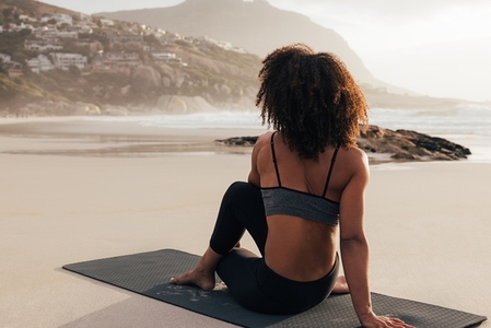 Back view of a young female practicing yoga at sunset  Woman in fitness wear sitting on a mat looking at hills on a shore