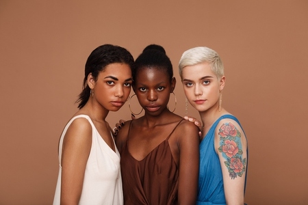 Group of three women of different races standing together against brown backdrop