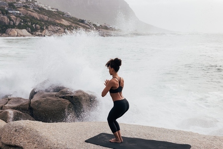 Woman practicing yoga while standing on stone against big waves and splashes  Female meditating at oceanside