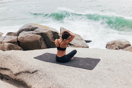 Rear view of young woman sitting on mat and warming up her neck by ocean  Female doing yoga and looking on waves