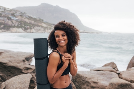 Portrait of a beautiful smiling woman standing with yoga mat by seaside  Female in sportswear relaxing after yoga looking away