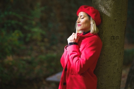 Gentle woman in red beret and coat leaning on tree