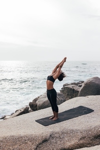 Young woman in fitness wear doing warming up exercises while standing on mat by seaside  Female practicing Hasta Uttanasana pose outdoors
