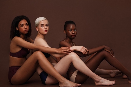 Side view of three diverse women sitting together in lingerie on brown background and looking at camera
