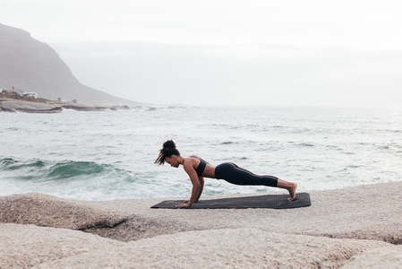 Side view of young woman in plank pose exercising on mat by ocean at evening