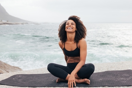 Laughing woman with curly hair sitting on mat by ocean  Female relaxing during yoga training outdoors