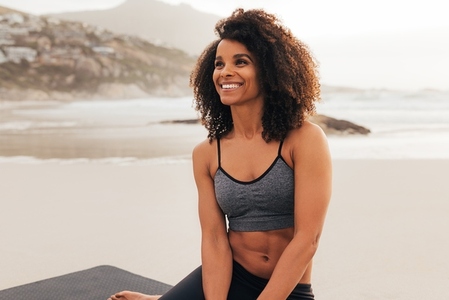 Portrait of a beautiful young woman with curly hair wearing sportswear sitting on beach relaxing at sunset