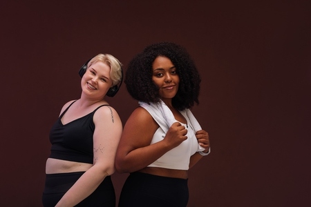 Two plus size woman standing back to back against brown background and looking at camera in studio