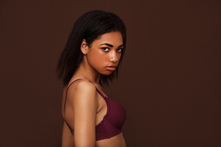 Side view of a young woman with black hair looking at camera  Young slim female in lingerie posing on brown background