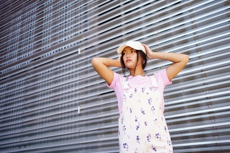 Low angle of glad Asian female in casual clothes  wearing cap  standing near modern building