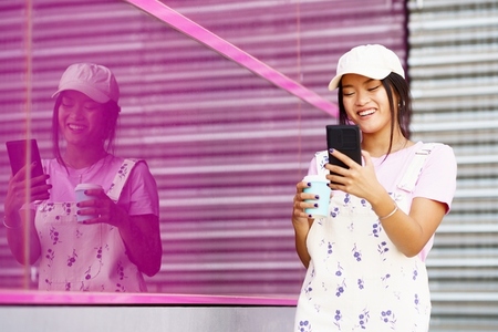 Asian female walking down the street  near a modern pink building  drinking coffee in a take away cup  while checking her smartphone