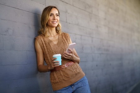 Middle aged woman taking a coffee break  Caucasian female using a paper cup and carrying her smartphone in her hand