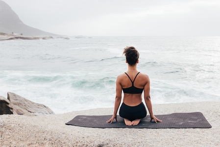 Woman sitting on a yoga mat and looking at ocean  Rear view of fit female sitting in Vajrasana pose outdoors