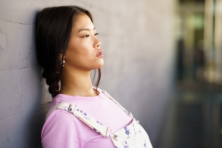 Young Chinese woman with a lost look and a serious expression leaning against an urban wall