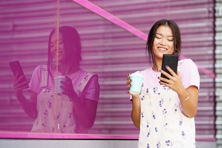 Asian girl near a modern pink building  drinking coffee in a take away cup  checking her smartphone
