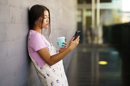 Asian female student taking a coffee break while consulting her smartphone