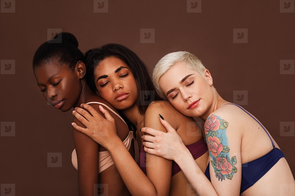 Three women in lingerie with closed eyes against a brown backdrop. Diverse females are embracing together