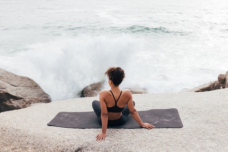Back view of female in fitness wear looking on waves while sitting on a yoga mat