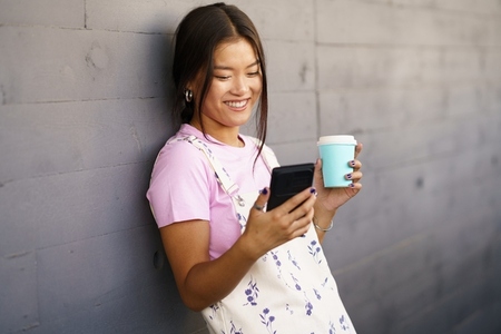 Cheerful Asian woman with coffee and smartphone leaning on wall
