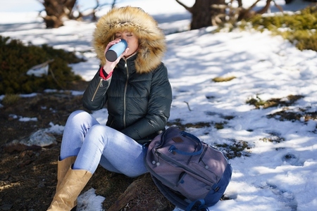 Woman drinking something hot from a metal thermos bottle sitting on a rock in the snowy mountains