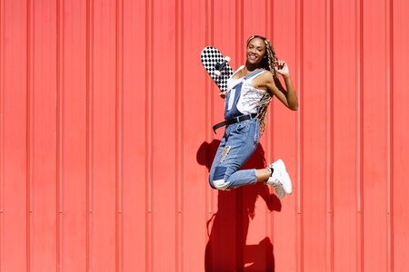 Black woman dressed casual  wtih a skateboard jumping with happiness on red urban wall background