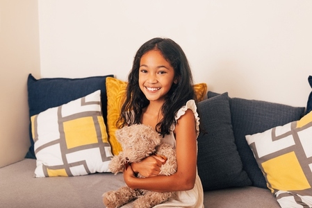 Smiling girl holding teddy bear while sitting on sofa looking at camera