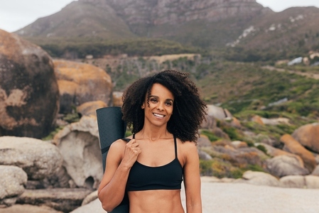 Portrait of a woman with curly hair with yoga mat standing outdoors by mountains