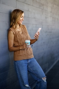 Cheerful woman with smartphone and coffee leaning on wall
