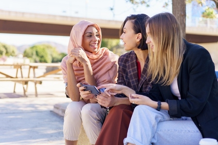 Positive multiethnic girlfriends sharing smartphone while resting on bench in park