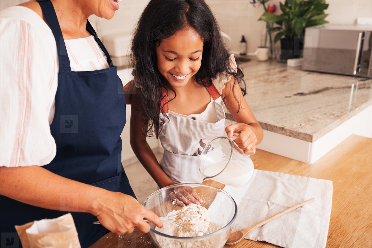 Smiling little girl and her grandma mixing flour in a bowl. Granddaughter pouring milk while granny whisking the flour
