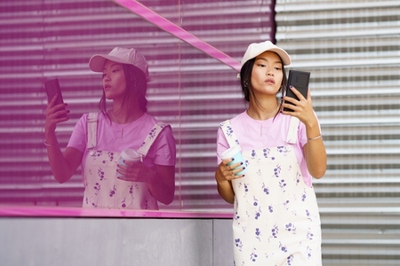 Young Asian woman with smartphone and coffee near pink wall