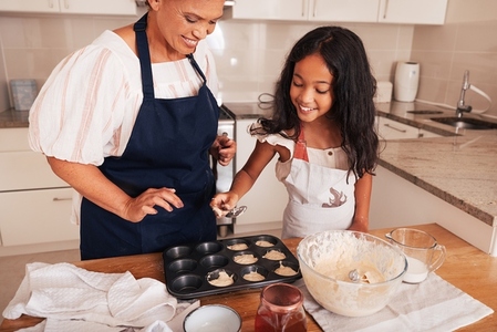 Girl in an apron pouring batter into cupcake molds while standing in the kitchen with grandma