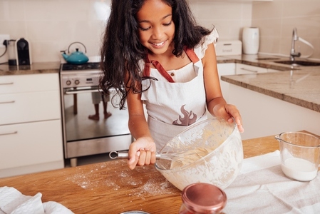 Smiling girl in apron mixing dough while standing in the kitchen