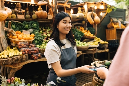 Outdoor market owner receiving payment from the buyer  Asian woman in an apron holding a pos terminal looking at a customer at a local farmer market