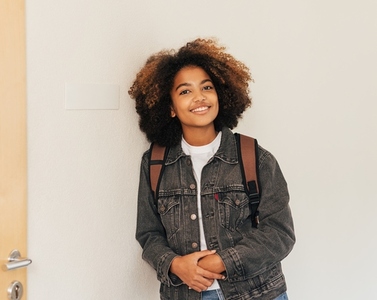 Portrait of a smiling girl with backpack leaning on wall in school looking at camera