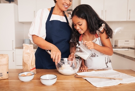Granny and granddaughter cook together  Smiling girl pouring milk while grandma mixing flour
