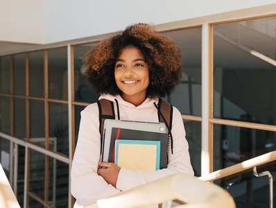 Smiling teenage girl with books and laptop standing on stairs in college and looking away
