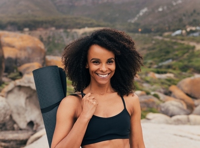 Fit woman with curly hair holding a yoga mat while standing outdoors after training